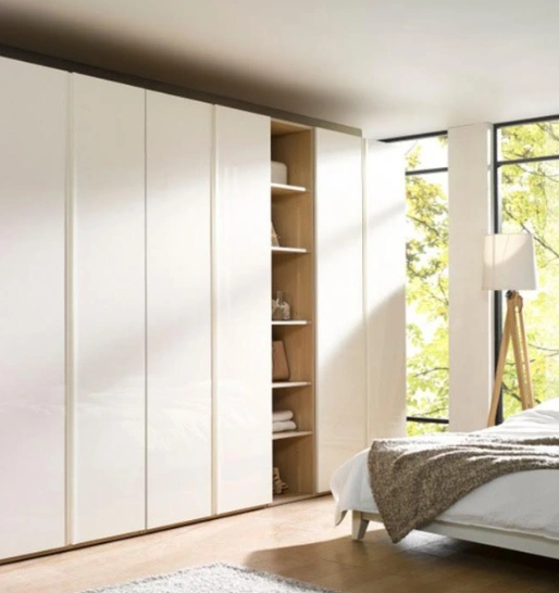 Modern bedroom with sliding wardrobe doors partially open revealing shelves, beside a bright window.