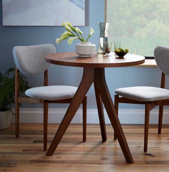 The room has blue walls, a large window, a round wooden dining table with a tripod base, chairs & vase.