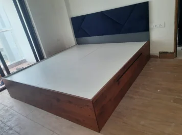 A sleek minimalist wooden platform bed with a stylish blue headboard, set in a serene, empty room.