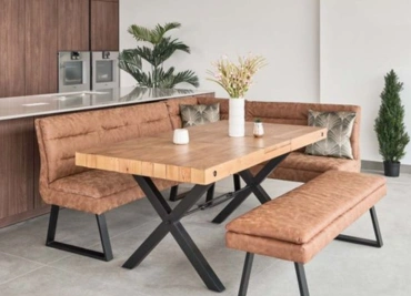 A modern dining area with a wooden table, tan corner bench seating, and greenery in the background. 