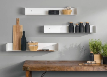 A Modern living in white shelves holding wooden bowls, black bottles & green plants on a wooden table